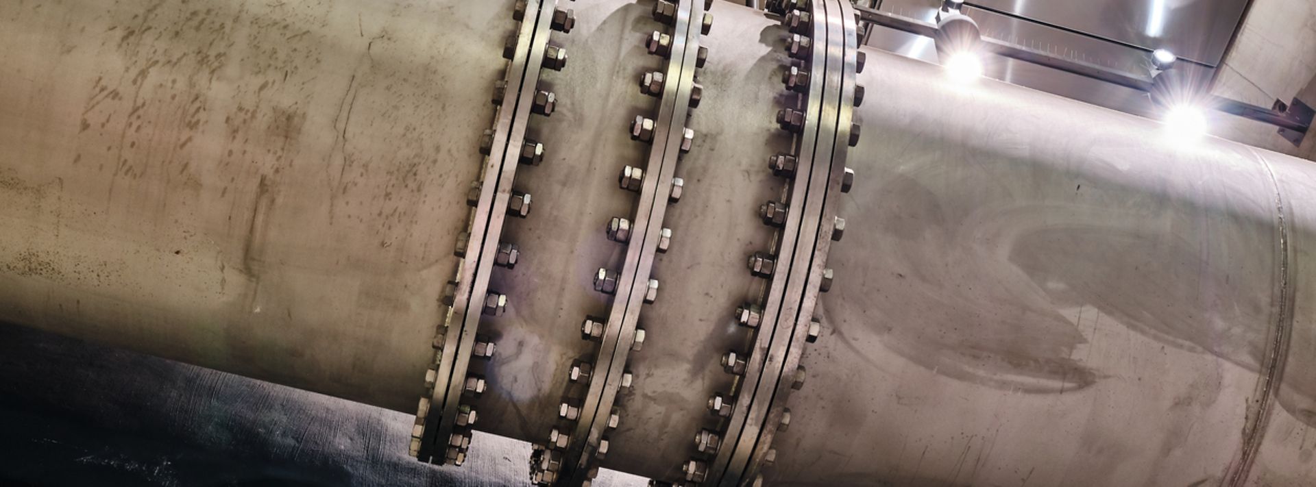 Großes Metallrohr mit mehreren Flanschen und Schraubverbindungen in einer beleuchteten Anlage