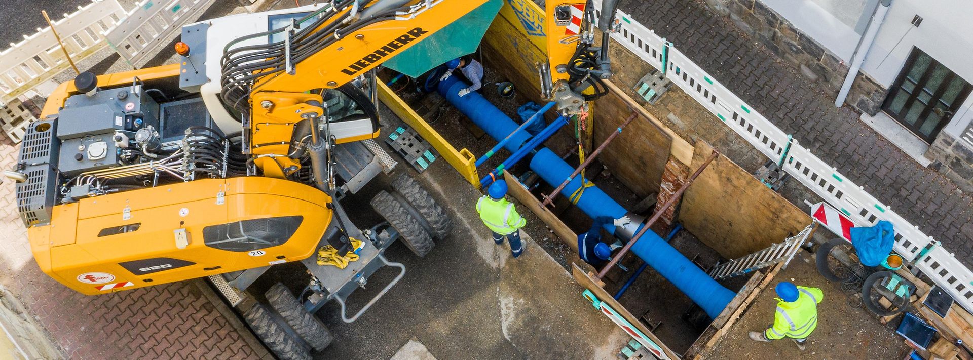 Eine Wasserleitung wird im Erdreich eingezogen. Ein Bagger und 2 Menschen stehen an der Baugrube.
