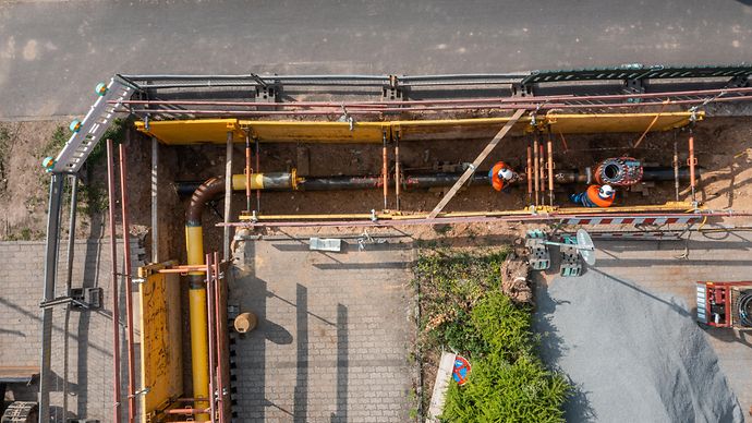 Baugrube mit freigelegter Rohrleitung; zwei Personen arbeiten im gesicherten Graben an Leitungen