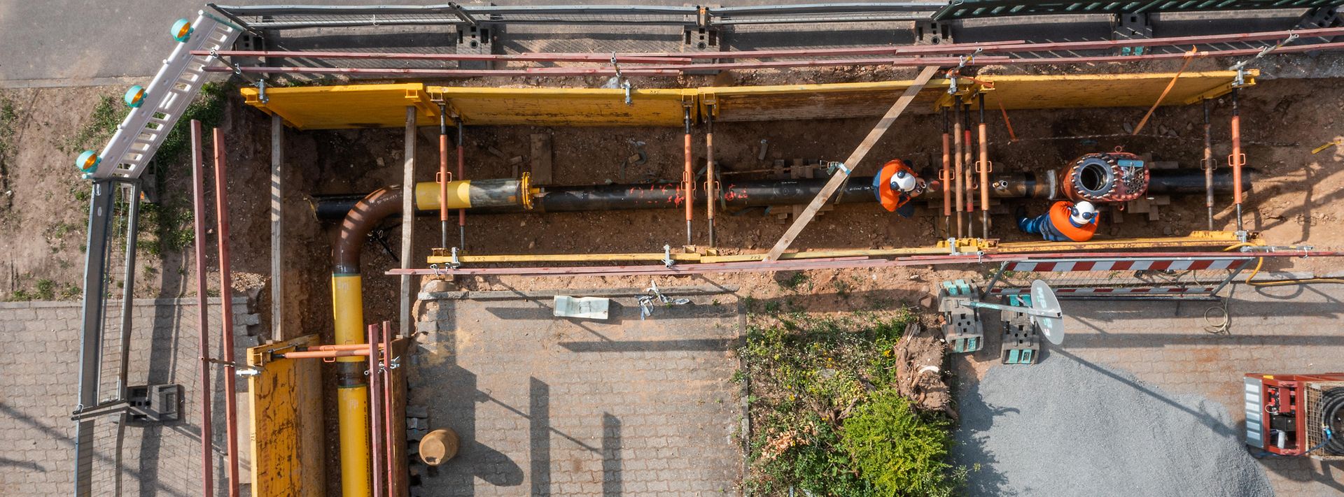 Baugrube mit freigelegter Rohrleitung; zwei Personen arbeiten im gesicherten Graben an Leitungen