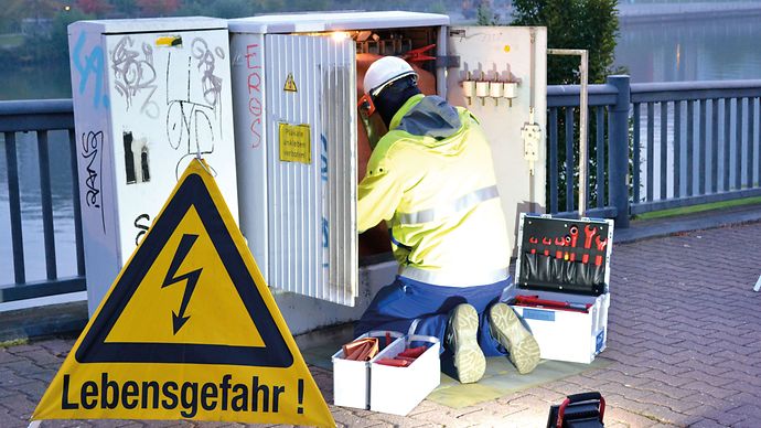 Ein Arbeiter in Schutzkleidung repariert einen offenen Stromkasten, daneben ein Werkzeugkasten und ein Warnschild.