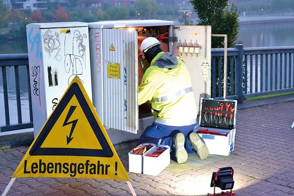 Ein Arbeiter in Schutzkleidung repariert einen offenen Stromkasten, daneben ein Werkzeugkasten und ein Warnschild.