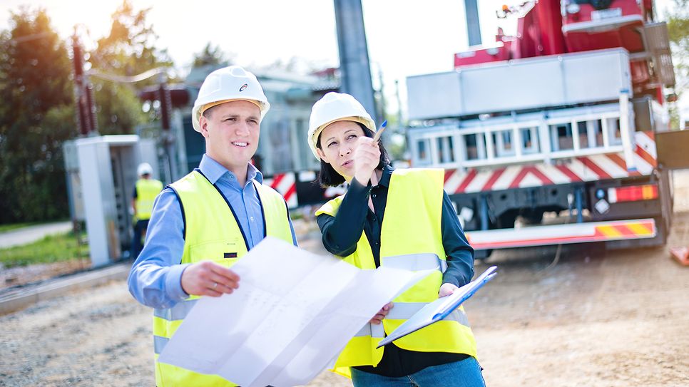 Zwei Mitarbeiter mit Helm, Warnweste und einem Plan in der Hand unterhalten sich 